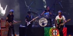 O guitarrista e vocalista Tim, o baterista Kalú, o guitarrista João Cabeleira e o guitarrista convidado Tó Trips em palco, com uma bandeira de Portugal.