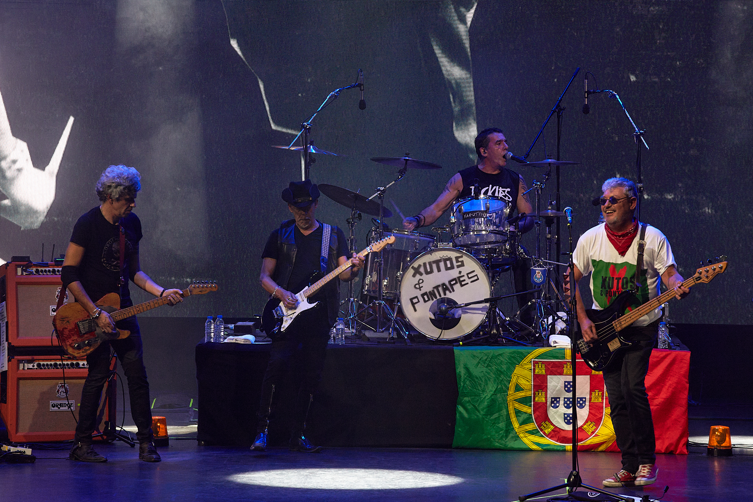 O guitarrista e vocalista Tim, o baterista Kalú, o guitarrista João Cabeleira e o guitarrista convidado Tó Trips em palco, com uma bandeira de Portugal.
